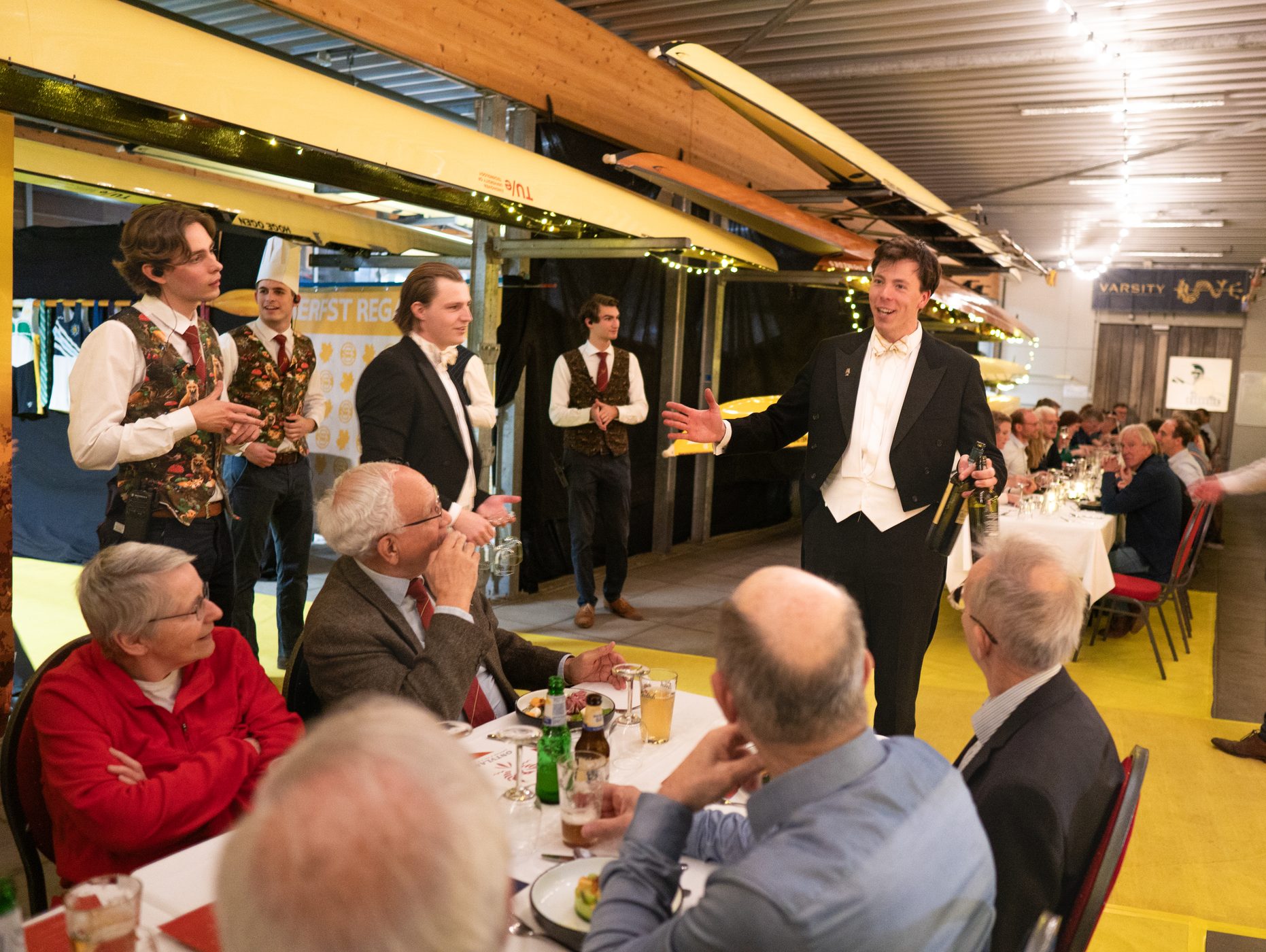 De vorige editie: een gastheer in rokkostuum spreekt de gasten aan tafel toe in de roeiloods, met de bediening en de kok achter hem. Opent het fotoalbum van de vorige editie.